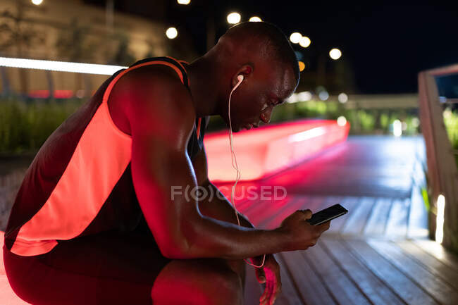 Vue latérale du coureur noir avec écouteurs utilisant le téléphone pendant l'entraînement la nuit — Photo de stock
