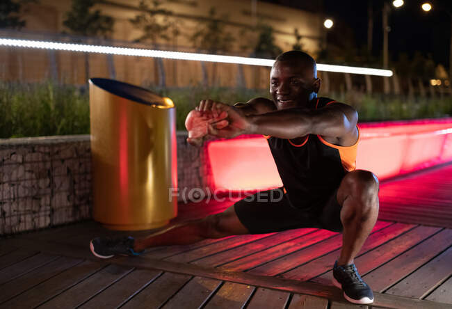 Happy black runner smiling and lunging during running workout at night in city — Stock Photo