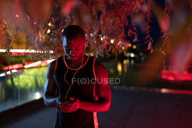 Ethnic runner browsing smartphone and listening to music under tree at night — Stock Photo