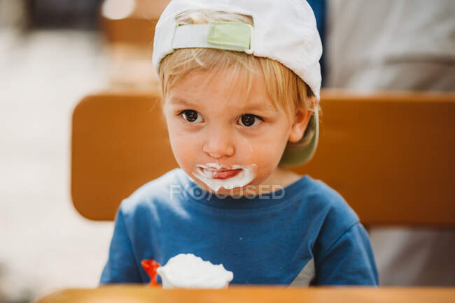 Niño blanco pequeño comiendo helado con la boca sucia y la tapa - foto de stock