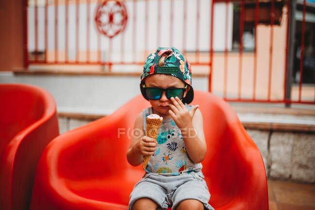 Белый мальчик ест мороженое в солнцезащитных очках и кепке к спине — стоковое фото