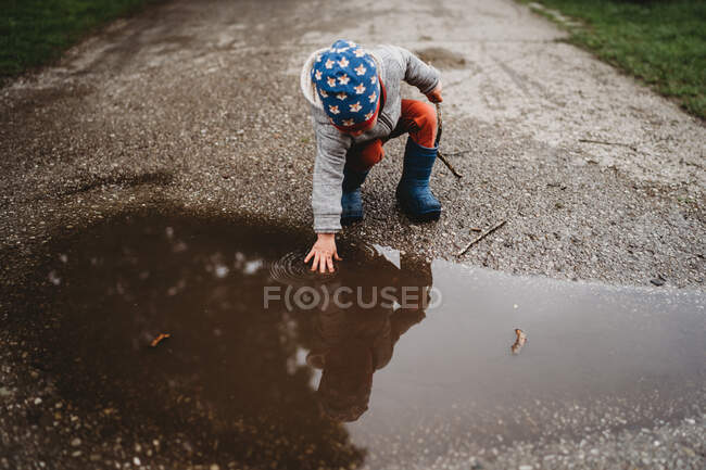 Niño varón joven poniendo la mano en un charco en el parque en el día nublado - foto de stock