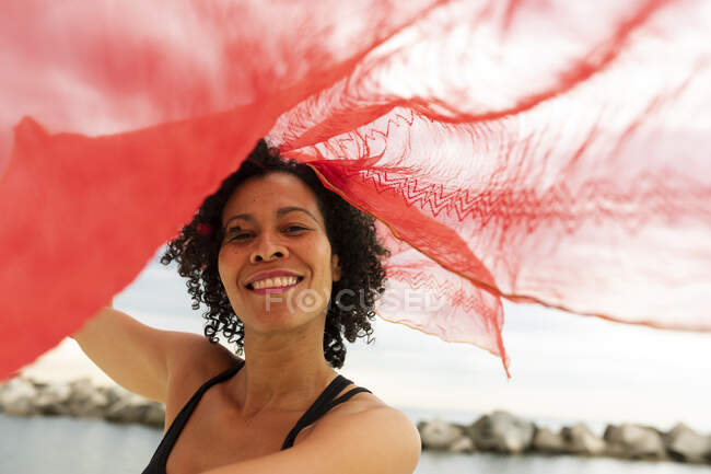 Pelo rizado africana americana mujer ondeando una bufanda roja en la playa - foto de stock