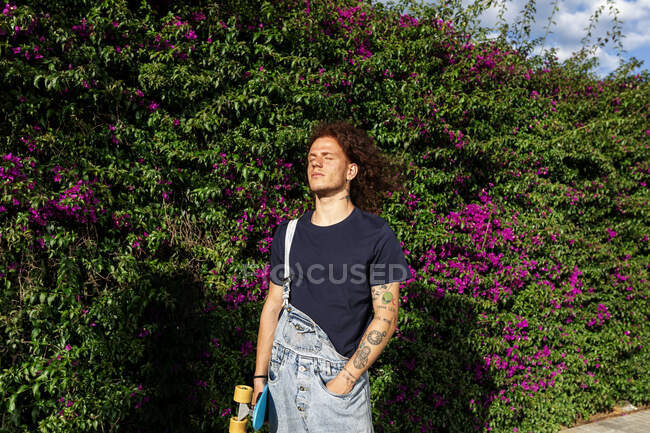 Кудрявые рыжие мужчины с татуировкой на волосах со скейтбордом на стене растений — стоковое фото