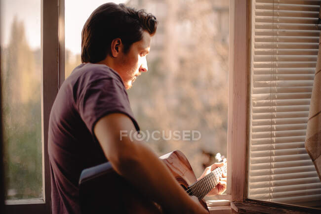 Joven tocando la guitarra mientras mira por la ventana mientras está de pie en el balcón en casa durante el auto aislamiento - foto de stock