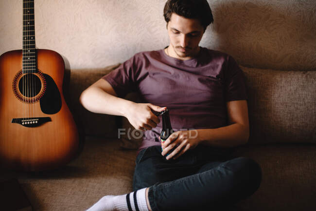 Молодий чоловік відкриває пляшку пива, сидячи на дивані вдома — стокове фото