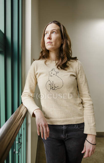 Una donna con un'espressione seria si appoggia su una rotaia che guarda fuori da una finestra — Foto stock