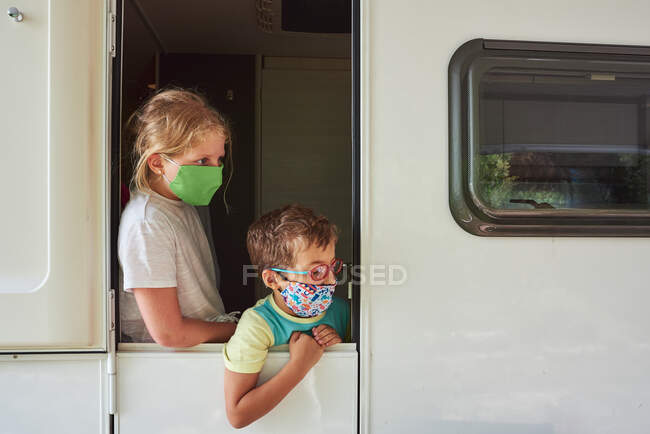 Hermanos de vacaciones en una caravana. Están en la puerta con una máscara de protección contra el virus corona - foto de stock