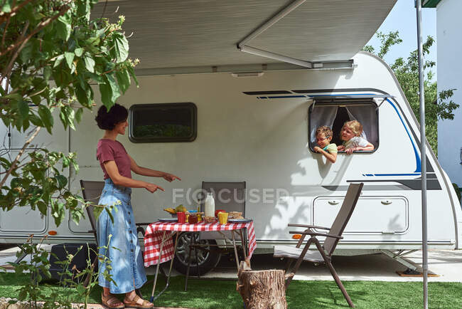 Deux enfants regardant par la fenêtre d'une caravane à un délicieux petit déjeuner. Leur mère les attend dehors avec le petit déjeuner prêt. — Photo de stock