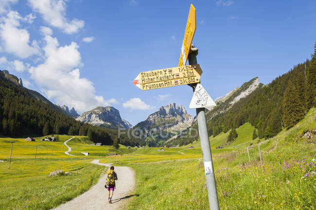 A woman hikes up a dirt road past a trail sign in Smtisertal, a valley in Alpstein,  Switzerland. — Stock Photo