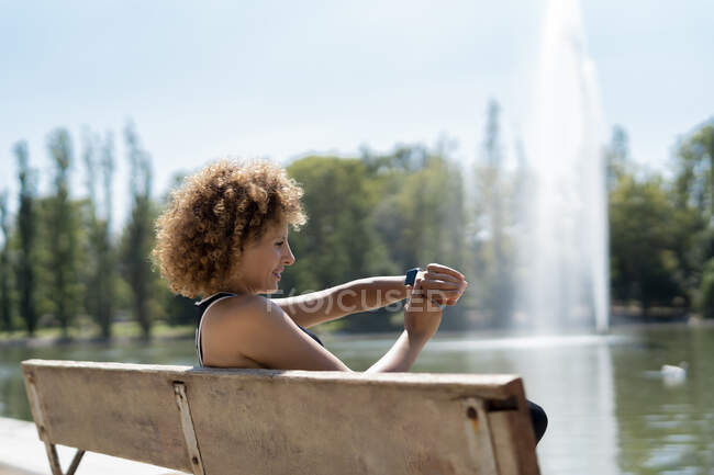 Mujer en un banco junto al lago usando su reloj inteligente - foto de stock