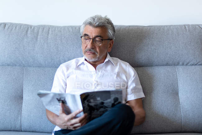 Older man with gray hair and glasses relaxing on his gray couch at home reading a magazine. — Stock Photo