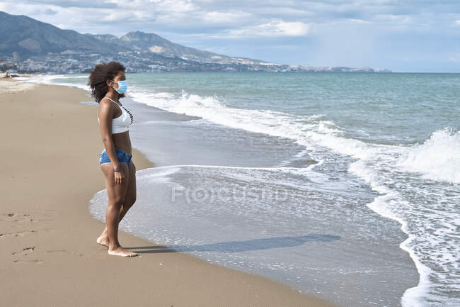 Mulher afro-americana caminha na praia com uma máscara — Fotografia de Stock