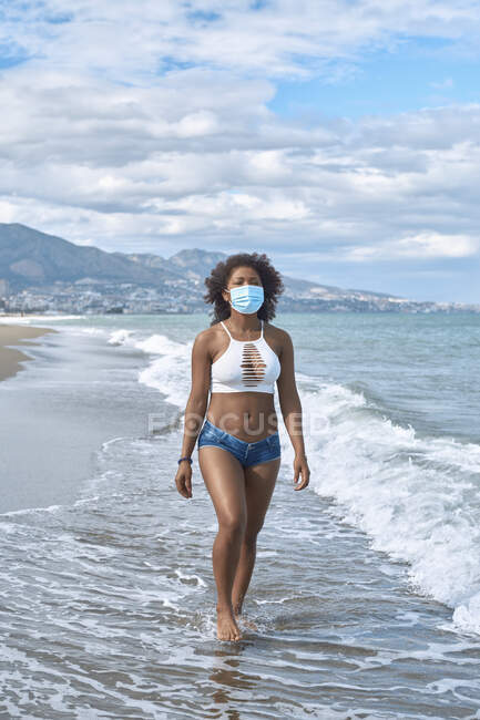 Mulher afro-americana caminha na praia com uma máscara — Fotografia de Stock
