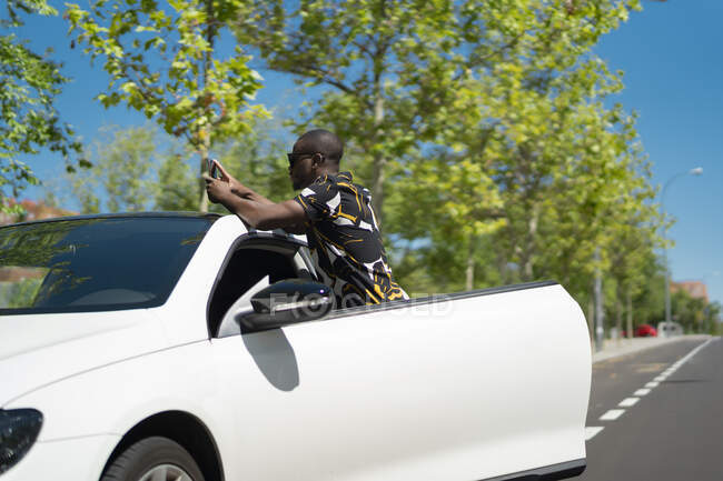 Молодой африканский мужчина фотографирует со своим смартфоном рядом со своей машиной — стоковое фото