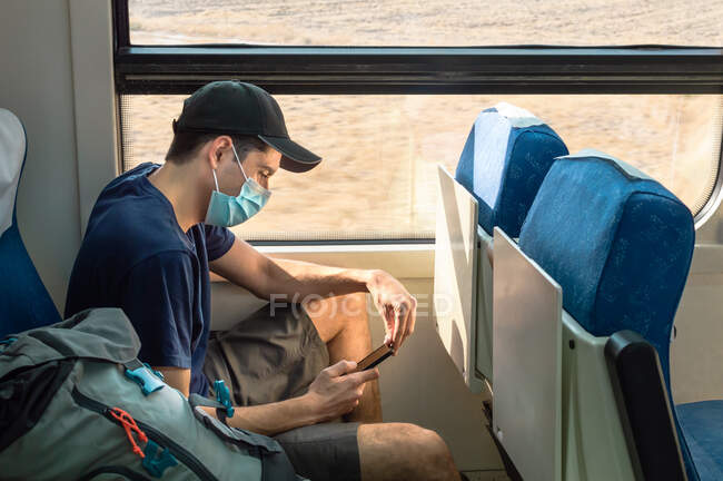 Mochilero con máscara facial usando teléfono móvil mientras viaja en tren. - foto de stock
