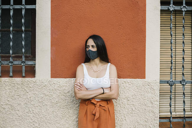 Porträt einer nachdenklichen Frau mit verschränkten Armen, die Gesichtsmaske an der Wand trägt — Stockfoto