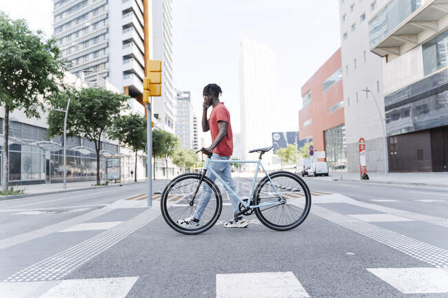 Side view of young ethnic male crossing street with bike and talking on smartphone — Stock Photo