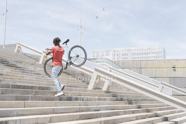 Задний вид черного человека, несущего велосипед и идущего вверх по лестнице по улице — стоковое фото