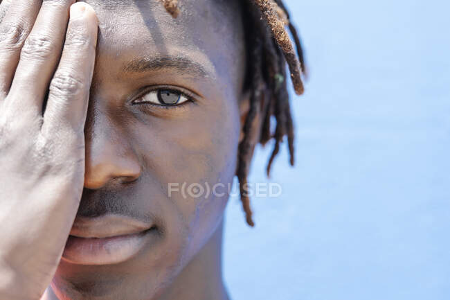 Crop young ethnic guy covering face and looking at camera against blue sky — Stock Photo