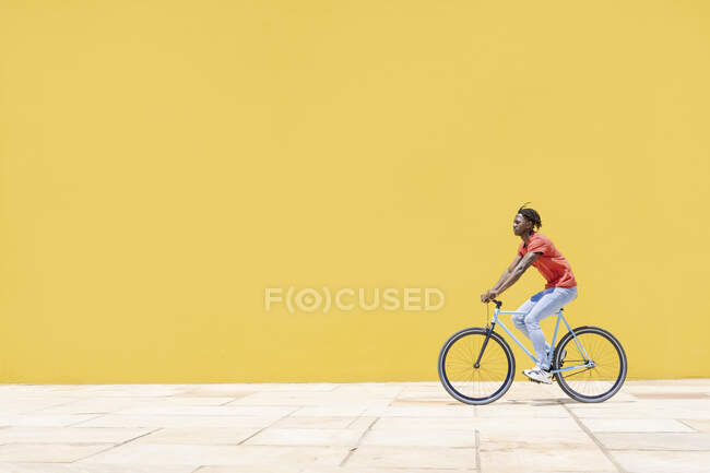 Vista lateral do homem étnico andar de bicicleta no pavimento perto da parede amarela na cidade — Fotografia de Stock