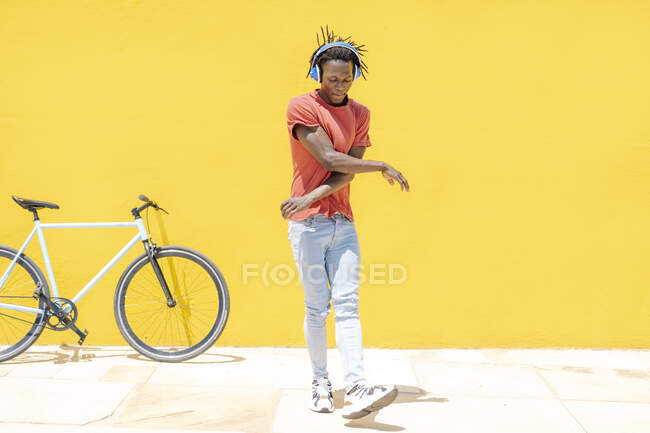 Joven hombre étnico escuchando música y bailando cerca de la bicicleta contra la pared amarilla - foto de stock