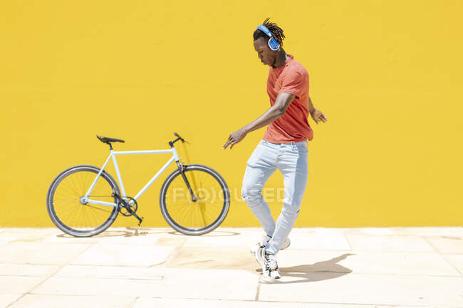Corpo inteiro cara preto em fones de ouvido dançando no caminho perto de bicicleta e parede amarela — Fotografia de Stock