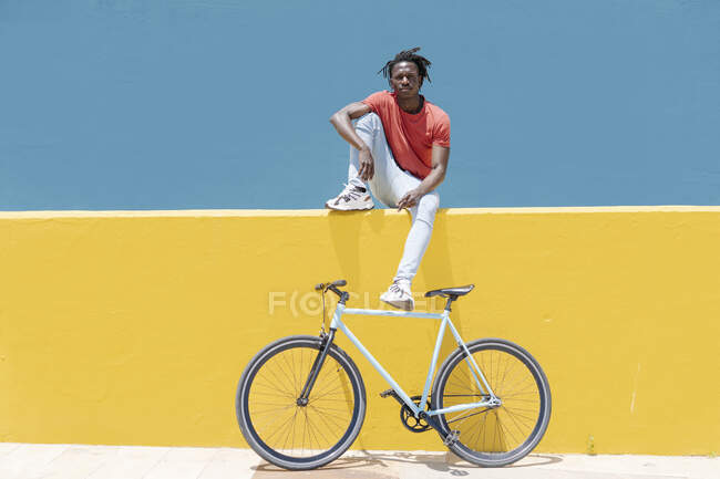 Young black guy sitting on yellow wall near bike against blue sky in city — Stock Photo