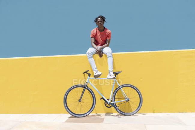 Joven negro sentado en la pared amarilla cerca de la bicicleta contra el cielo azul en la ciudad - foto de stock