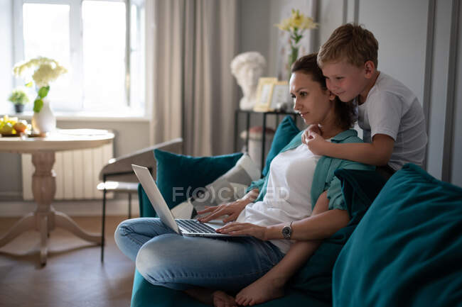 Menino sorrindo e abraçando a mãe usando laptop para o trabalho no sofá — Fotografia de Stock