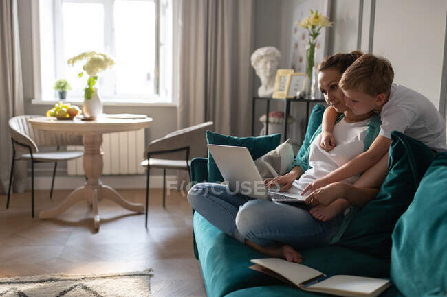 Kleiner Junge umarmt Mutter mit Laptop und hilft bei Remote-Projekt auf Sofa — Stockfoto
