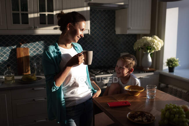 Mãe encantada com caneca e pequeno filho sorrindo e tomando café da manhã juntos — Fotografia de Stock