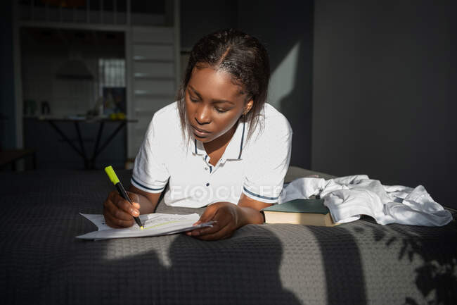 Mujer negra haciendo marcas en el documento mientras está acostada en la cama y estudiando en casa - foto de stock