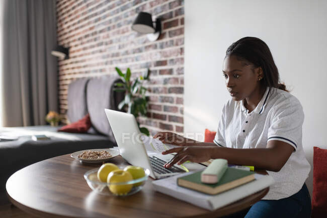 Молодая черная женщина с бумагами просматривает данные на ноутбуке во время учебы дома — стоковое фото