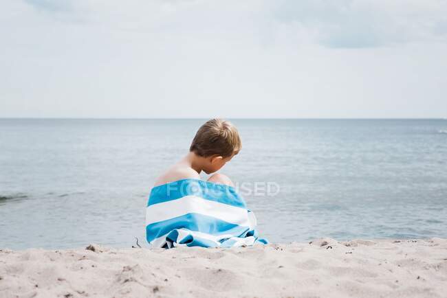 Jeune garçon assis enveloppé dans une serviette rayée seul sur la plage — Photo de stock