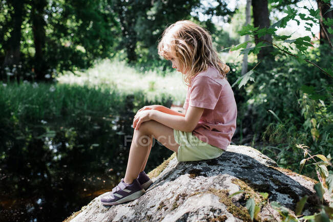 Молода дівчина виглядає сумно і самотньо на скелі біля річки влітку — стокове фото
