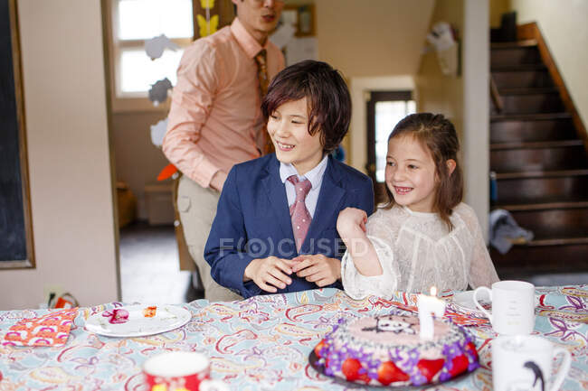 Los hermanos sonrientes se sientan a la mesa en ropa elegante con pastel de cumpleaños encendido - foto de stock