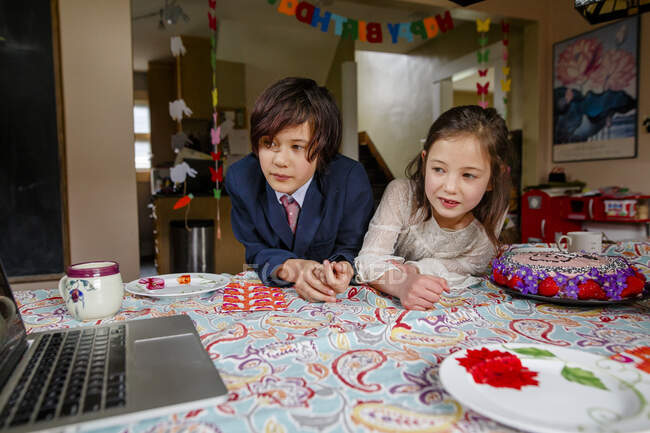 Un ragazzo e una ragazza si siedono a tavola con la torta di compleanno guardando un computer — Foto stock