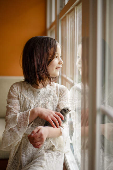 Fille heureuse regardant par la fenêtre avec bébé poussin niché dans ses bras — Photo de stock