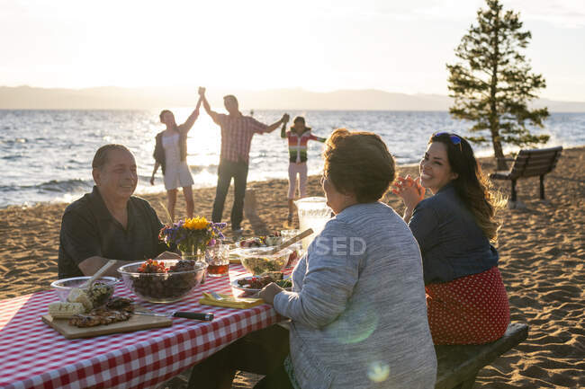 Mutter und Großeltern unterhalten sich beim Abendessen, während der Vater und seine Töchter bei Sonnenuntergang am Nevada Beach in Lake Tahoe, Nevada, im Sand spielen. — Stockfoto