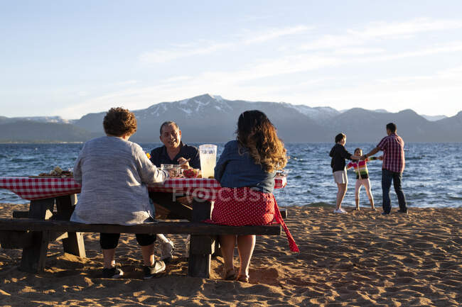 Mutter und Großeltern unterhalten sich beim Abendessen, während der Vater und seine Töchter bei Sonnenuntergang am Nevada Beach in Lake Tahoe, Nevada, im Sand spielen. — Stockfoto