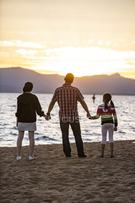 Отец держит своих дочерей за руки, пока они смотрят закат на Невада-Бич в озере Тахо, Невада. — стоковое фото