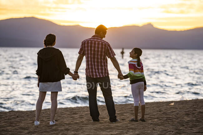 Un padre se toma de la mano con sus hijas mientras observan la puesta de sol en Nevada Beach en Lake Tahoe, Nevada. - foto de stock