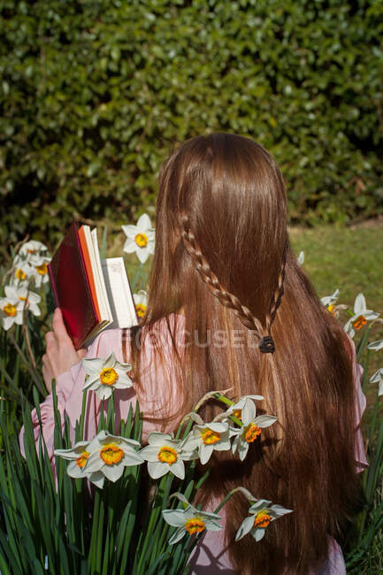 Visão traseira da menina de cabelos castanhos lendo um livro sentado entre as flores — Fotografia de Stock