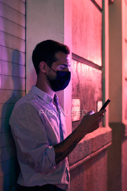 Jovem com uma máscara olha para seu telefone celular à noite — Fotografia de Stock