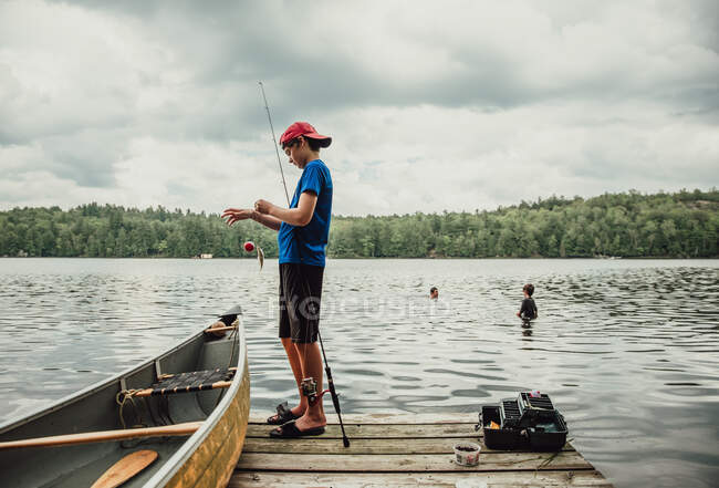 Adolescente menino pesca a partir de uma doca em um lago com irmãos nadando nas proximidades. — Fotografia de Stock