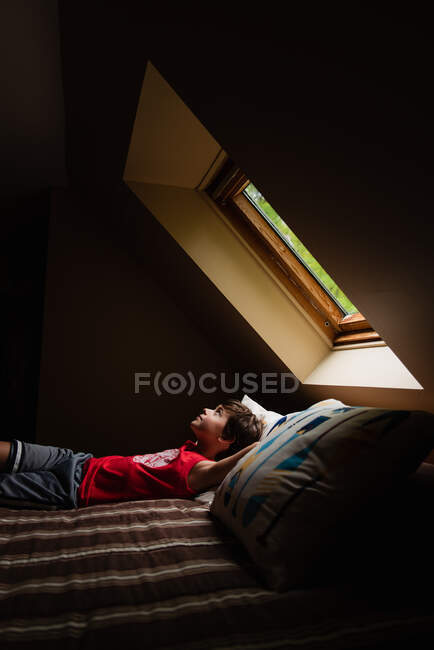 Jeune garçon allongé sur le lit regardant à travers une lumière du ciel dans une pièce sombre. — Photo de stock