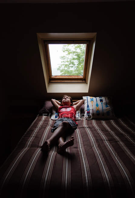 Kleiner Junge liegt auf Bett und schaut durch ein Himmelslicht in einem dunklen Raum nach oben. — Stockfoto