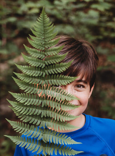 Porträt eines kleinen lächelnden Jungen, der sein halbes Gesicht mit Farnblatt bedeckt. — Stockfoto