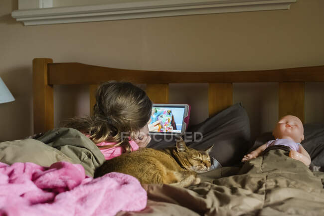 Young girl watching her device in bed with her cat and doll — Stock Photo